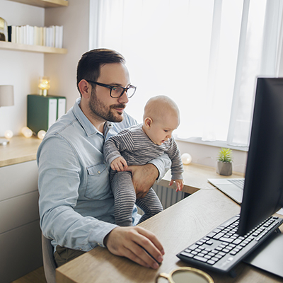 Photo of man sitting in front of computer with a baby on his lap.